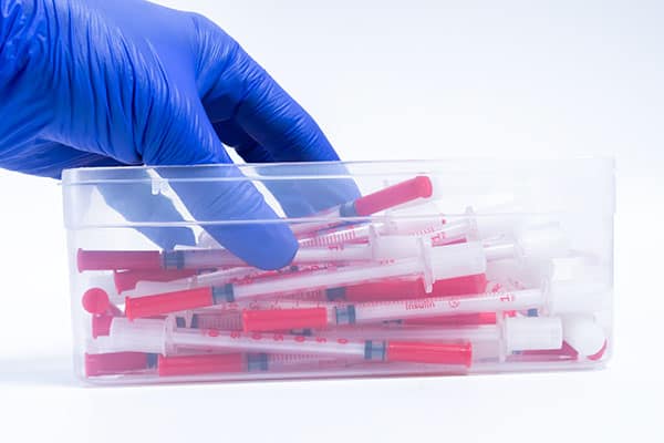 Blue gloved hand reaching into sharps container full of syringes